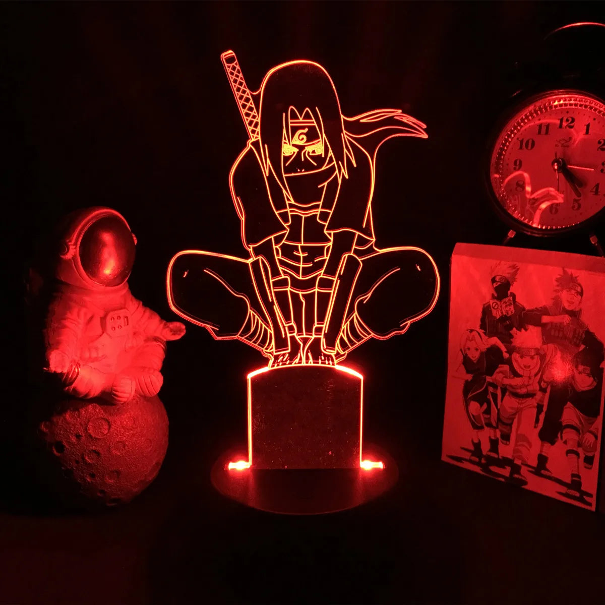 <img src="itachi-on-pole-nightlight.jpg" alt="Red LED lamp depicting Itachi Uchiha from Naruto in a crouching pose, just before the Uchiha clan massacre. On the left, a glowing astronaut figurine, and on the right, a clock and an image featuring other Naruto characters, creating a moody, dimly lit decor.">